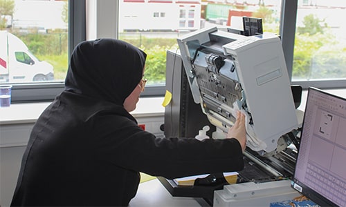 Fatima, an employee wearing a headscarf, operates a large scanner in a bright office. She works carefully while inspecting the machine.