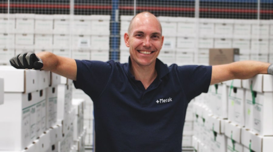 A smiling man in a blue Merak work polo casually leans against stacks of boxes in a warehouse, conveying an organized and professional work environment.