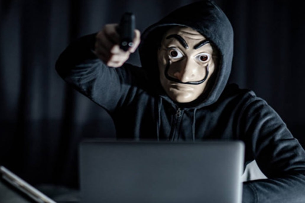 Een hacker zit met een masker achter een computer met een geweer in zijn hand
